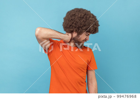 Portrait of man with Afro hairstyle in T-shirt touching neck, feeling acute pain moving and turning head, suffering spine problems, osteochondrosis. Indoor studio shot isolated on blue background. Portrait of man with Afro hairstyle in T-shirt touching neck, feeling acute pain moving and turning head, suffering spine problems, osteochondrosis. Indoor studio shot isolated on blue background. 99476968