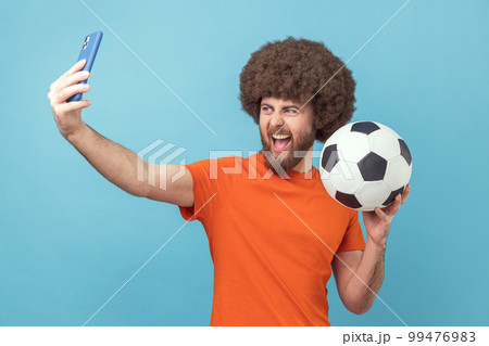Portrait of excited man with Afro hairstyle wearing orange T-shirt standing with soccer ball and broadcasting livestream from football match. Indoor studio shot isolated on blue background. 99476983