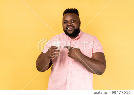 Portrait of smiling satisfied bearded man wearing pink shirt holding glass of water with ice, pointing at cold beverage, looking at camera. Indoor studio shot isolated on yellow background. Portrait of smiling satisfied bearded man wearing pink shirt holding glass of water with ice, pointing at cold beverage, looking at camera. Indoor studio shot isolated on yellow background. 99477016