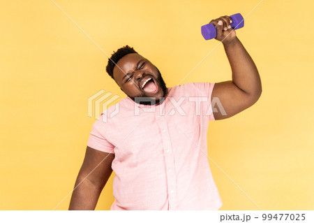 Portrait of funny positive bearded man wearing pink shirt standing and raised arm with dumbbell, making effort raising heavy sport equipment. Indoor studio shot isolated on yellow background. Portrait of funny positive bearded man wearing pink shirt standing and raised arm with dumbbell, making effort raising heavy sport equipment. Indoor studio shot isolated on yellow background. 99477025