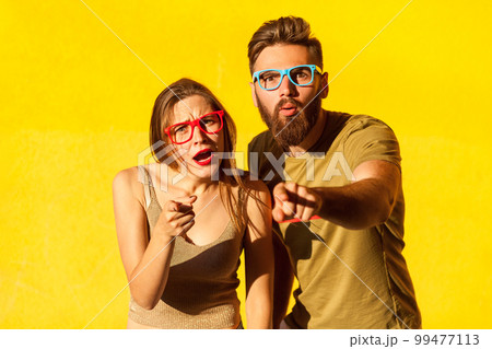 Portrait of astonished amazed man and woman in glasses standing with shocked face and pointing to camera, having surprised expression. Indoor studio shot isolated on yellow background. 99477113