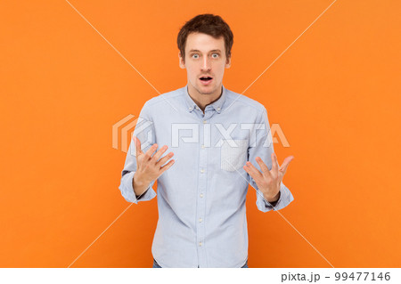 Portrait of shocked unhappy young adult man standing with raised hands, asking what, misunderstanding, wearing light blue shirt. Indoor studio shot isolated on orange background. 99477146