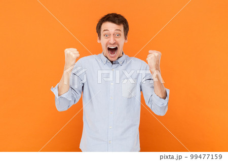 Portrait of extremely happy man standing with clenched fists, celebrating his success, screaming hurray, wearing light blue shirt. Indoor studio shot isolated on orange background. Portrait of extremely happy man standing with clenched fists, celebrating his success, screaming hurray, wearing light blue shirt. Indoor studio shot isolated on orange background. 99477159