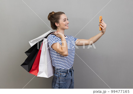 Side view portrait of woman wearing striped T-shirt broadcasting livestream and boasting her purchase, holding shopping, using smartphone. Indoor studio shot isolated on gray background. 99477165