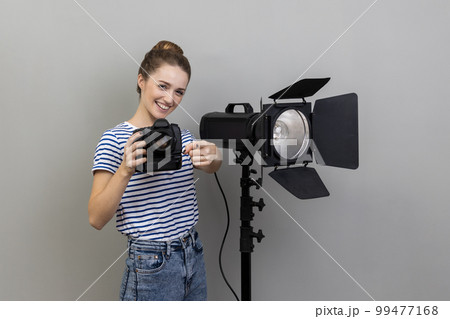 Portrait of smiling satisfied woman photographer photographing, holding photo camera and using illumination equipment, pointing to camera. Indoor studio shot isolated on gray background. 99477168