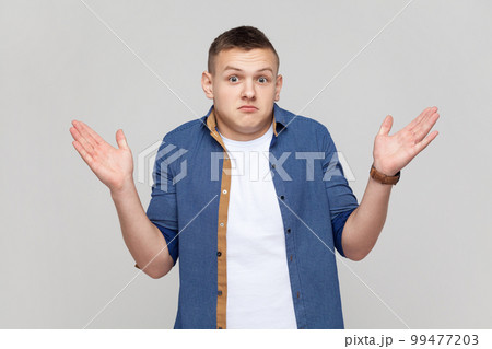 I don't know, who cares. Portrait of confused clueless teenager boy wearing blue shirt shrugging shoulders, making no idea gesture, whatever. Indoor studio shot isolated on gray background. 99477203