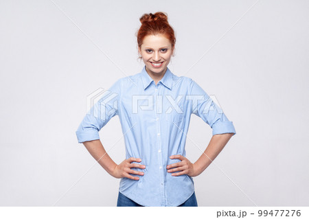 Portrait of positive happy pretty redhead woman wearing blue shirt standing with with hands on hips, looking at camera with toothy smile. Indoor studio shot isolated on gray background. 99477276