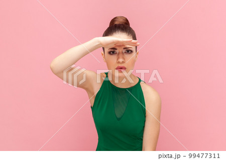 Portrait of serious young adult woman with bun hairstyle standing with hand near forehead and looking far at distance, wearing green dress. Indoor studio shot isolated on pink background. Portrait of serious young adult woman with bun hairstyle standing with hand near forehead and looking far at distance, wearing green dress. Indoor studio shot isolated on pink background. 99477311