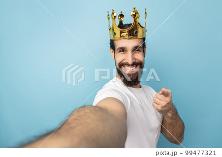Portrait of smiling man with beard wearing white T-shirt and gold crown, pointing at camera, POV, point of view of photo, expressing happiness. Indoor studio shot isolated on blue background. Portrait of smiling man with beard wearing white T-shirt and gold crown, pointing at camera, POV, point of view of photo, expressing happiness. Indoor studio shot isolated on blue background. 99477321