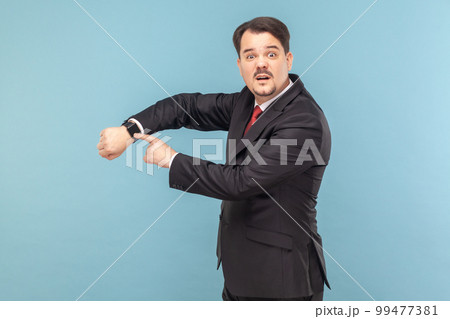 Confused man with mustache standing and looking at camera with puzzled expression, pointing at his wristwatch, wearing black suit with red tie. Indoor studio shot isolated on light blue background. 99477381