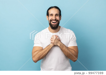 Portrait of satisfied delighted handsome man with beard wearing white T-shirt standing with hands together, looking at camera with amazed positive face. Indoor studio shot isolated on blue background. Portrait of satisfied delighted handsome man with beard wearing white T-shirt standing with hands together, looking at camera with amazed positive face. Indoor studio shot isolated on blue background. 99477399