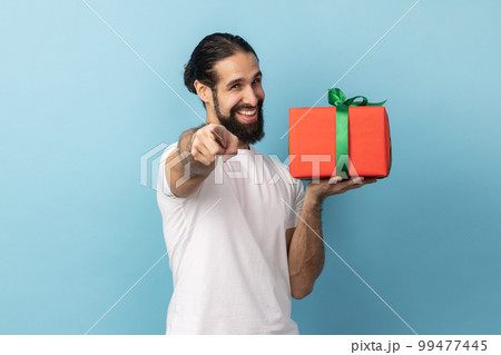 Portrait of positive man with beard wearing white T-shirt giving present and pointing finger to camera, sharing holiday gift, congratulating. Indoor studio shot isolated on blue background. 99477445