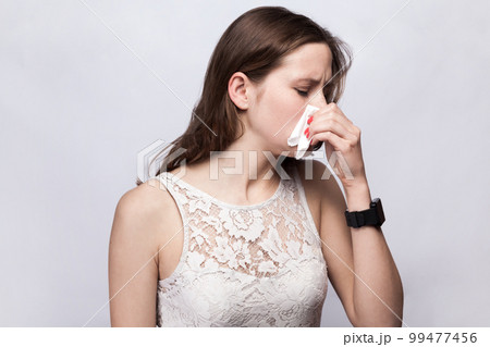 Portrait of unhealthy attractive young adult woman wearing white dress catching cold, having flu symptoms and runny nose, sneezing. Indoor studio shot isolated on gray background. Portrait of unhealthy attractive young adult woman wearing white dress catching cold, having flu symptoms and runny nose, sneezing. Indoor studio shot isolated on gray background. 99477456