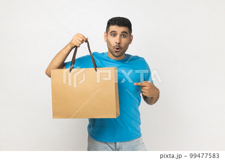 Portrait of shocked surprised astonished unshaven man wearing blue T- shirt standing pointing at shopping bag, great purchase in mall. Indoor studio shot isolated on gray background. 99477583