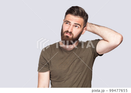 Portrait of pensive bearded man wearing dark green T-shirt touching head and thinking about important question, making hard decision. Indoor studio shot isolated on light gray background. 99477655