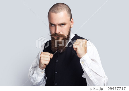 Portrait of serious angry man with beard and mustache standing with clenched fists, being ready to fight with somebody, looking at camera. Indoor studio shot isolated on gray background. Portrait of serious angry man with beard and mustache standing with clenched fists, being ready to fight with somebody, looking at camera. Indoor studio shot isolated on gray background. 99477717