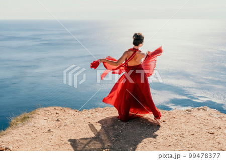 Woman in red dress on sea. Side view a Young beautiful sensual woman in a red long dress posing on a rock high above the sea on sunset. Girl on the nature on blue sky background. Fashion photo. 99478377