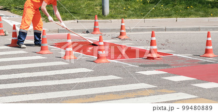Road repair. Man in orange work clothes repairs pedestrian crossing in morning, summer and spring. Road is fenced with special cones. Road repair. Man in orange work clothes repairs pedestrian crossing in morning, summer and spring. Road is fenced with special cones. 99480889