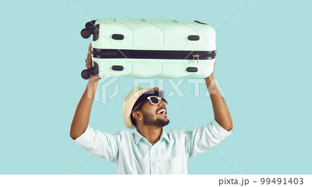 Happy Indian man having fun lifting travel suitcase above his head on light blue background. 99491403
