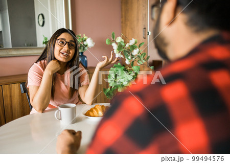 Happy couple eating breakfast and talking at dining table in morning. Indian girl and latin guy. Relationship and diversity concept Happy couple eating breakfast and talking at dining table in morning. Indian girl and latin guy. Relationship and diversity concept 99494576