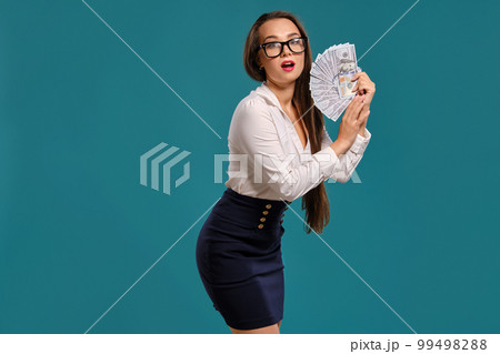 Brunette girl in glasses, wearing in a black short skirt and white blouse is posing holding a fan of hundred dollar bills against a blue background. 99498288