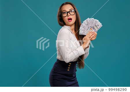 Brunette girl in glasses, wearing in a black short skirt and white blouse is posing holding a fan of hundred dollar bills against a blue background. 99498290