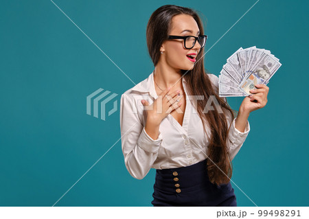 Brunette girl in glasses, wearing in a black short skirt and white blouse is posing holding a fan of hundred dollar bills against a blue background. 99498291