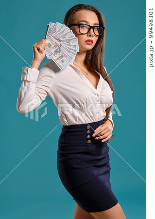 Brunette girl in glasses, wearing in a black short skirt and white blouse is posing holding a fan of hundred dollar bills against a blue background. Brunette girl in glasses, wearing in a black short skirt and white blouse is posing holding a fan of hundred dollar bills against a blue background. 99498301