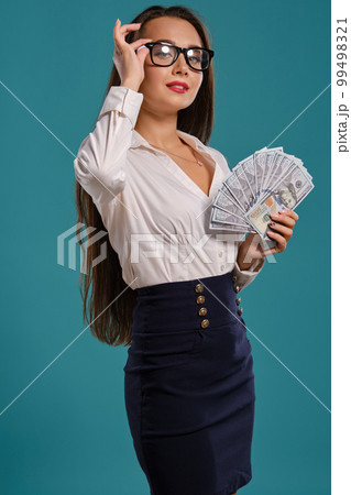 Brunette girl in glasses, wearing in a black short skirt and white blouse is posing holding a fan of hundred dollar bills against a blue background. 99498321