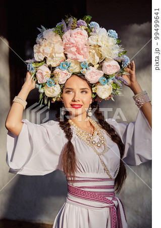 Brunette girl in a white ukrainian authentic national costume and a wreath of flowers is posing against a white hut. Close-up. 99499641