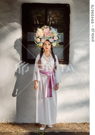 Brunette girl in a white ukrainian authentic national costume and a wreath of flowers is posing against a white hut. Close-up. Brunette girl in a white ukrainian authentic national costume and a wreath of flowers is posing against a white hut. Close-up. 99499676