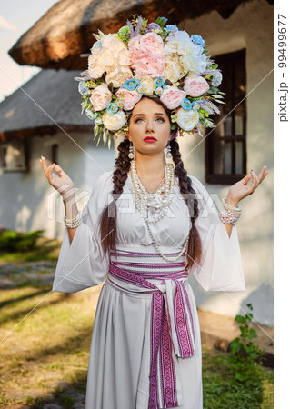 Brunette girl in a white ukrainian authentic national costume and a wreath of flowers is posing against a white hut. Close-up. 99499677