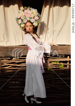 Brunette girl in a white ukrainian authentic national costume and a wreath of flowers is posing against a terrace. 99499717