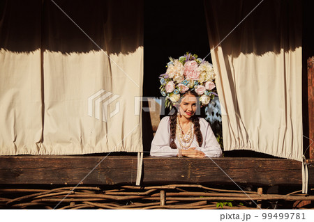 Brunette girl in a white ukrainian authentic national costume and a wreath of flowers is is looking out of the window. 99499781