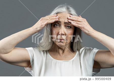 Sad old european lady with gray hair touches her face, wrinkles with hands, isolated on gray background 99500409