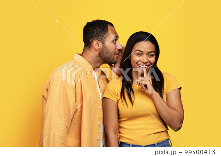Smiling young african american male whispers to lady ear, female puts finger to lips, making secret gesture 99500413