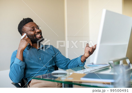 Excited black businessman talking on cellphone and looking at computer monitor, sitting at workplace in office Excited black businessman talking on cellphone and looking at computer monitor, sitting at workplace in office 99500491