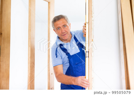 Concentrated male builder in uniform using a waterpass on the wooden door of the apartment 99505079