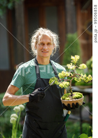 Blonde caucasian man planting flowers in pot with garden tools. Man wearing green t-shirt and black apron. Man spending peaceful morning in garden hear house. Blonde caucasian man planting flowers in pot with garden tools. Man wearing green t-shirt and black apron. Man spending peaceful morning in garden hear house. 99506858