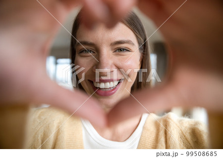 Cheerful beautiful young woman looking at camera through hand heart Cheerful beautiful young woman looking at camera through hand heart 99508685