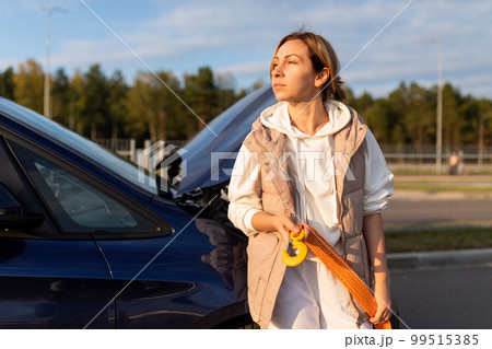 an adult Caucasian businesswoman with a cable for transporting a car stands at the raised hood of a 99515385