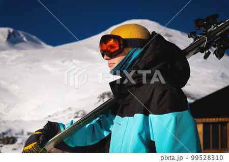 skier holds a pair of skis and looks at the snow-capped mountains to the side. A guy with skis on his shoulder stands in the mountains covered with snow. Man with winter gear having fun skier holds a pair of skis and looks at the snow-capped mountains to the side. A guy with skis on his shoulder stands in the mountains covered with snow. Man with winter gear having fun 99528310