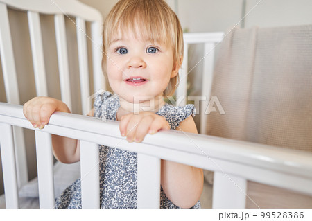 Happy baby girl one and half standing in the crib. Smiling 18 months child playing in sunny bedroom. Baby cot in the parent's bedroom Happy baby girl one and half standing in the crib. Smiling 18 months child playing in sunny bedroom. Baby cot in the parent's bedroom 99528386