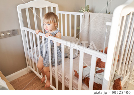 Happy baby girl one and half standing in the crib. Smiling 18 months child playing in sunny bedroom. Baby cot in the parent's bedroom Happy baby girl one and half standing in the crib. Smiling 18 months child playing in sunny bedroom. Baby cot in the parent's bedroom 99528400