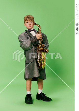 Stylish little school age boy in huge oversize retro style clothes and big male shoes posing with ukulele guitar isolated on green background. 99529087