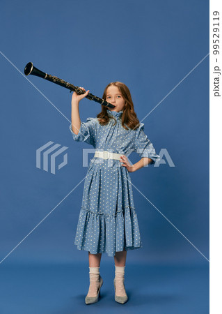 Monochrome portrait of charming nice little girl in dress and big shoes playing on clarinet over blue background. Music, fashion, hobby concept 99529119