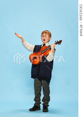 Happy kid, little musician wearing big size clothes posing with violin isolated over blue background. Education, music, classic art, new vision, fun concept 99529161
