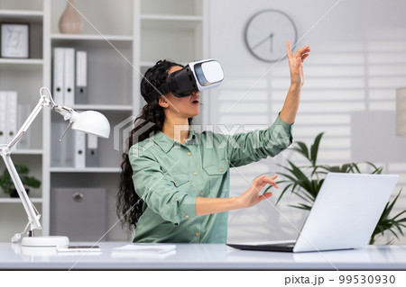 Young successful woman inside the office using vr glasses, virtual reality simulator cross charts and documents online, latin american woman using laptop at work. 99530930