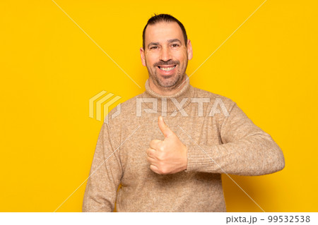 Bearded hispanic man wearing beige turtleneck posing with thumbs up in approval and self-confidence isolated over yellow background. Concept of success and positivism. 99532538
