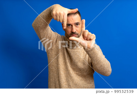 Handsome bearded happy male looks through a frame formed by his hands. An attractive man makes a frame with his fingers, isolated on a blue background with copy space for text. People concept. 99532539